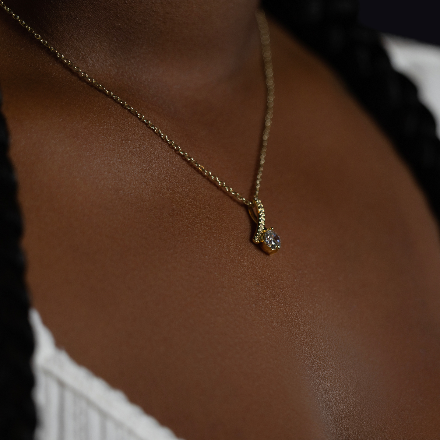 person wearing a gold chain necklace with a small silver pendant featuring a clear gemstone, shown against dark brown skin