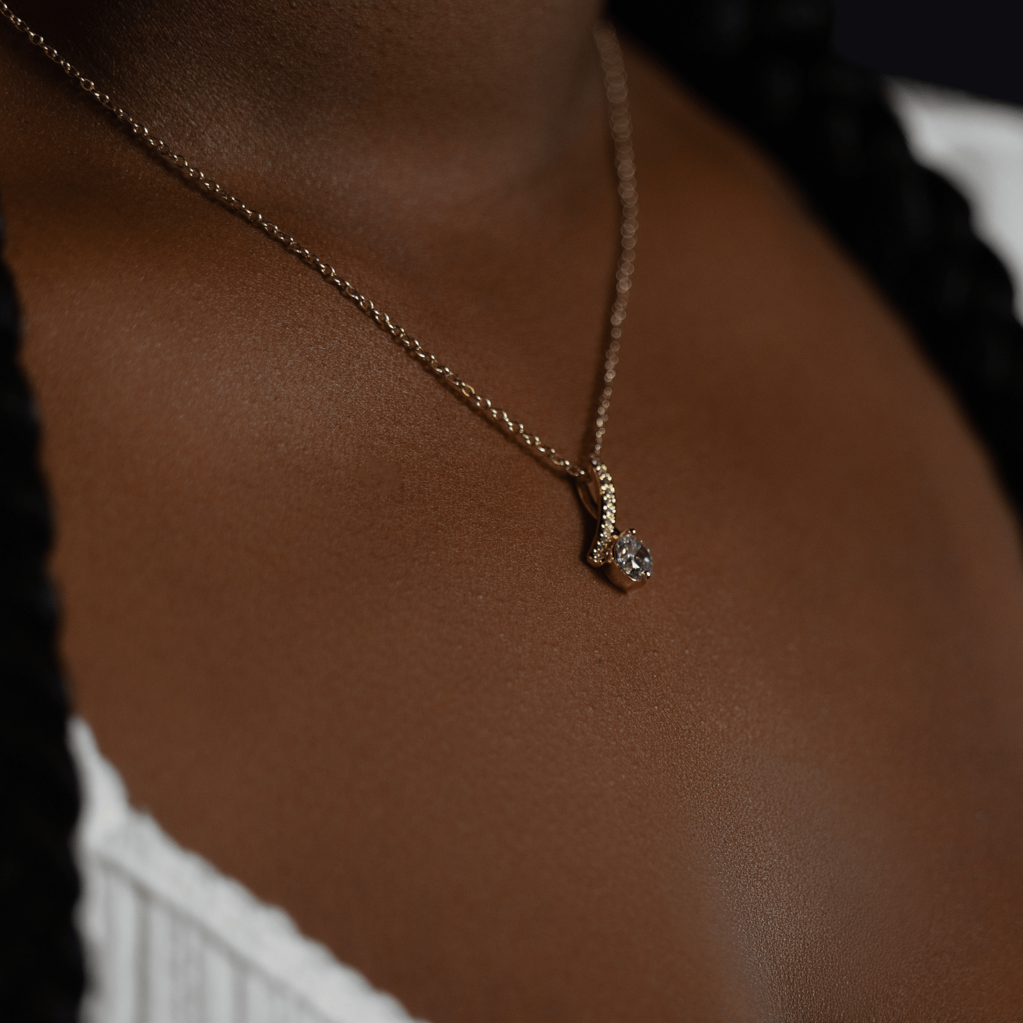 person wearing a gold chain necklace with a small silver pendant featuring a clear gemstone, shown against dark brown skin