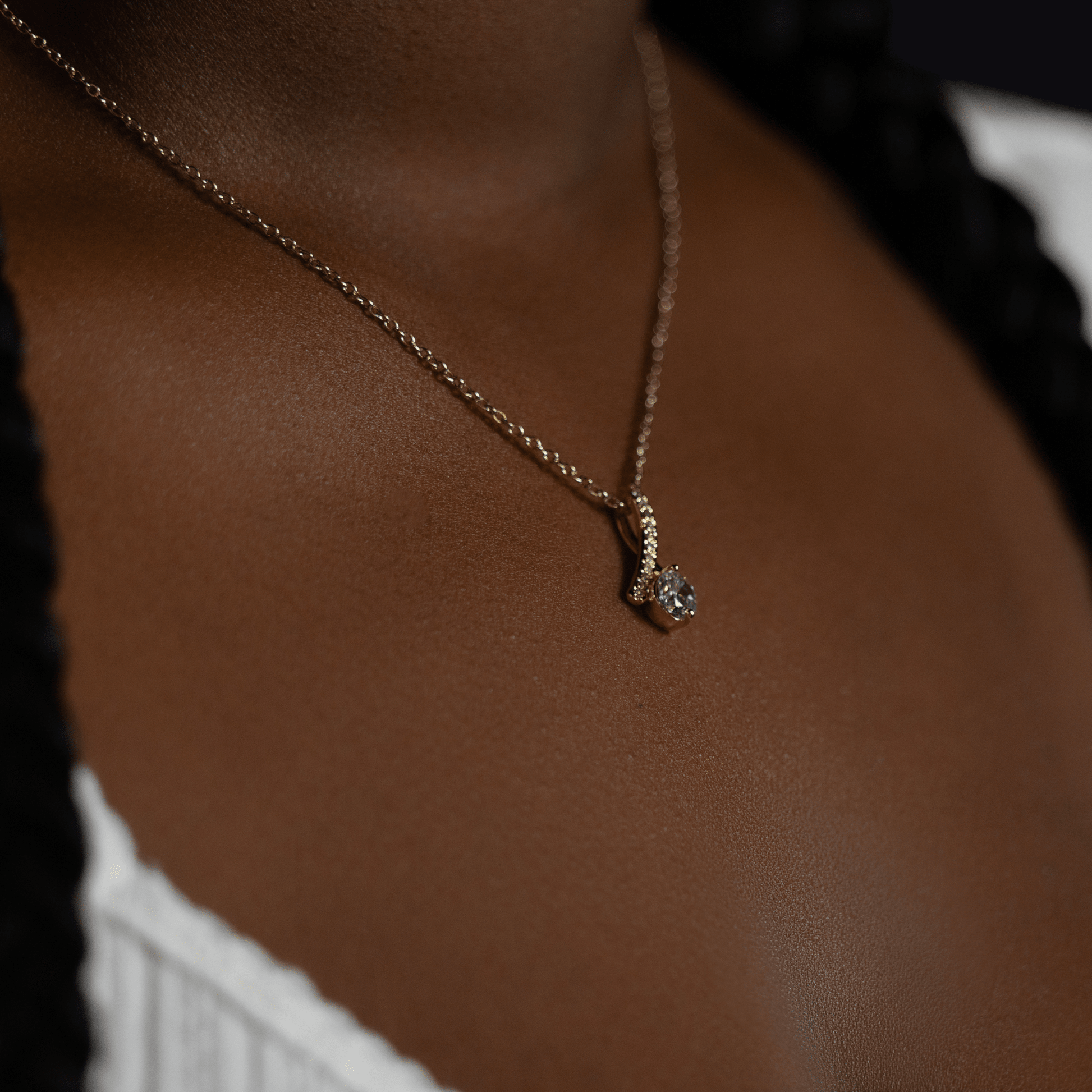 person wearing a gold chain necklace with a small silver pendant featuring a clear gemstone, shown against dark brown skin