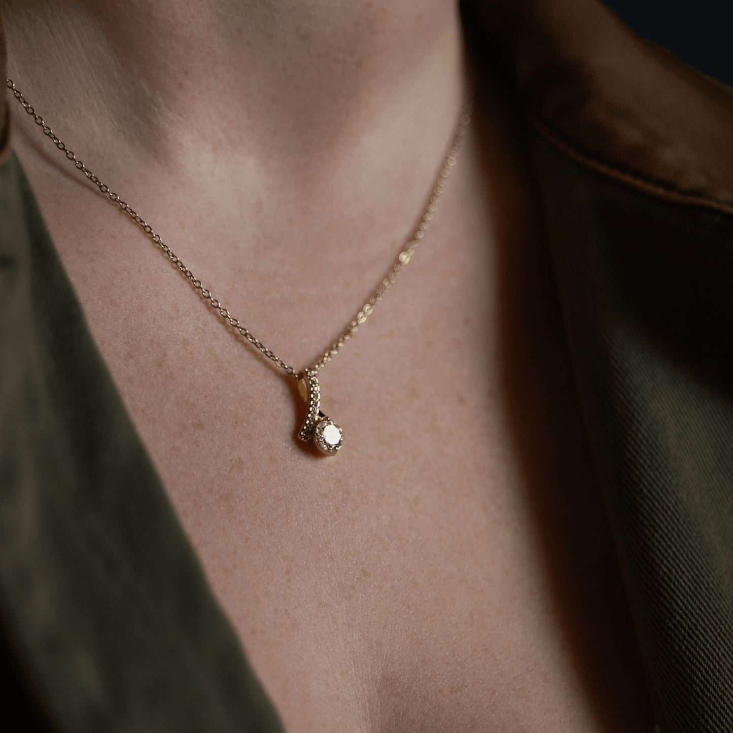 Gold chain necklace with a pendant featuring a clear gemstone, model in an olive green shirt over a white top.