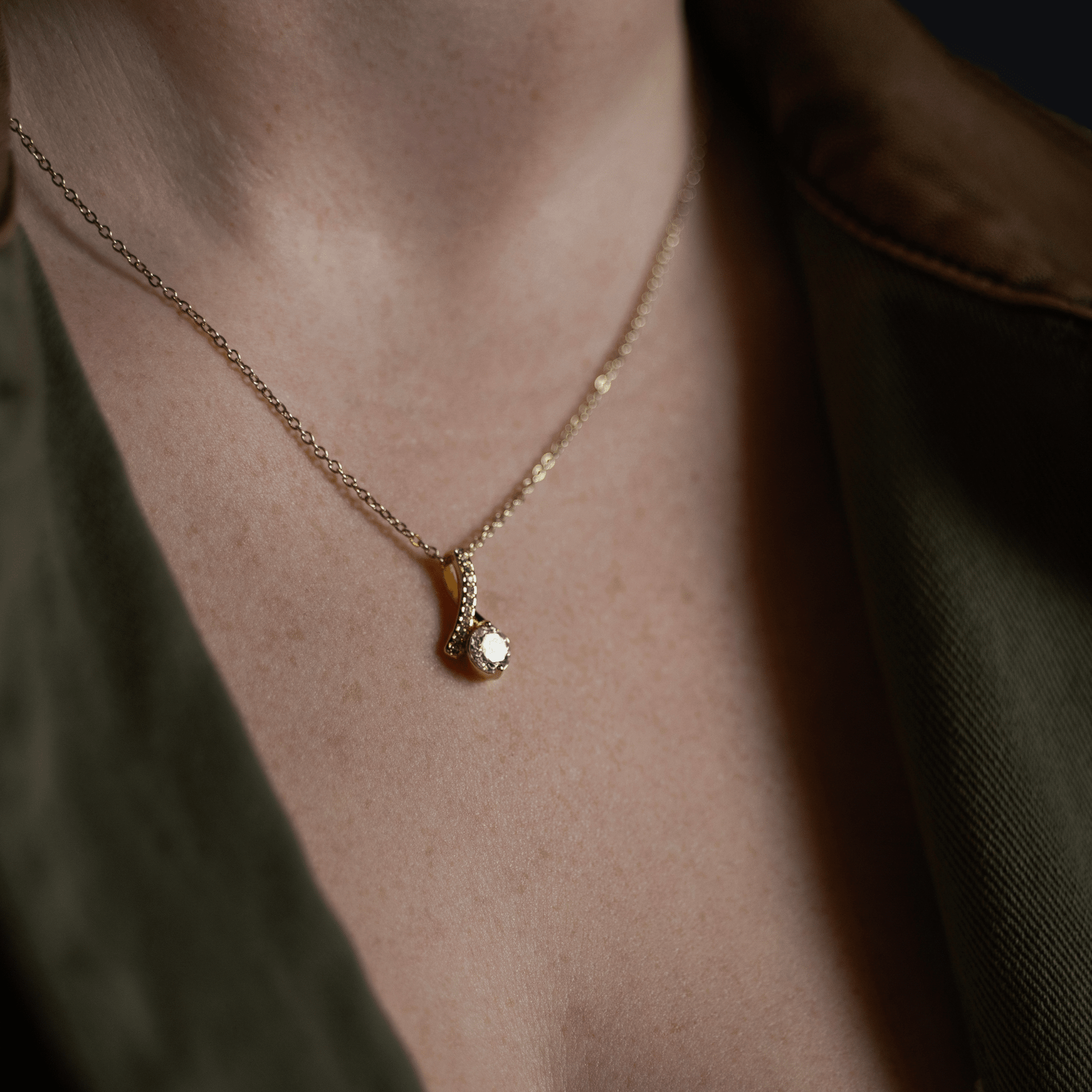 Gold chain necklace with a pendant featuring a clear gemstone, model in an olive green shirt over a white top.