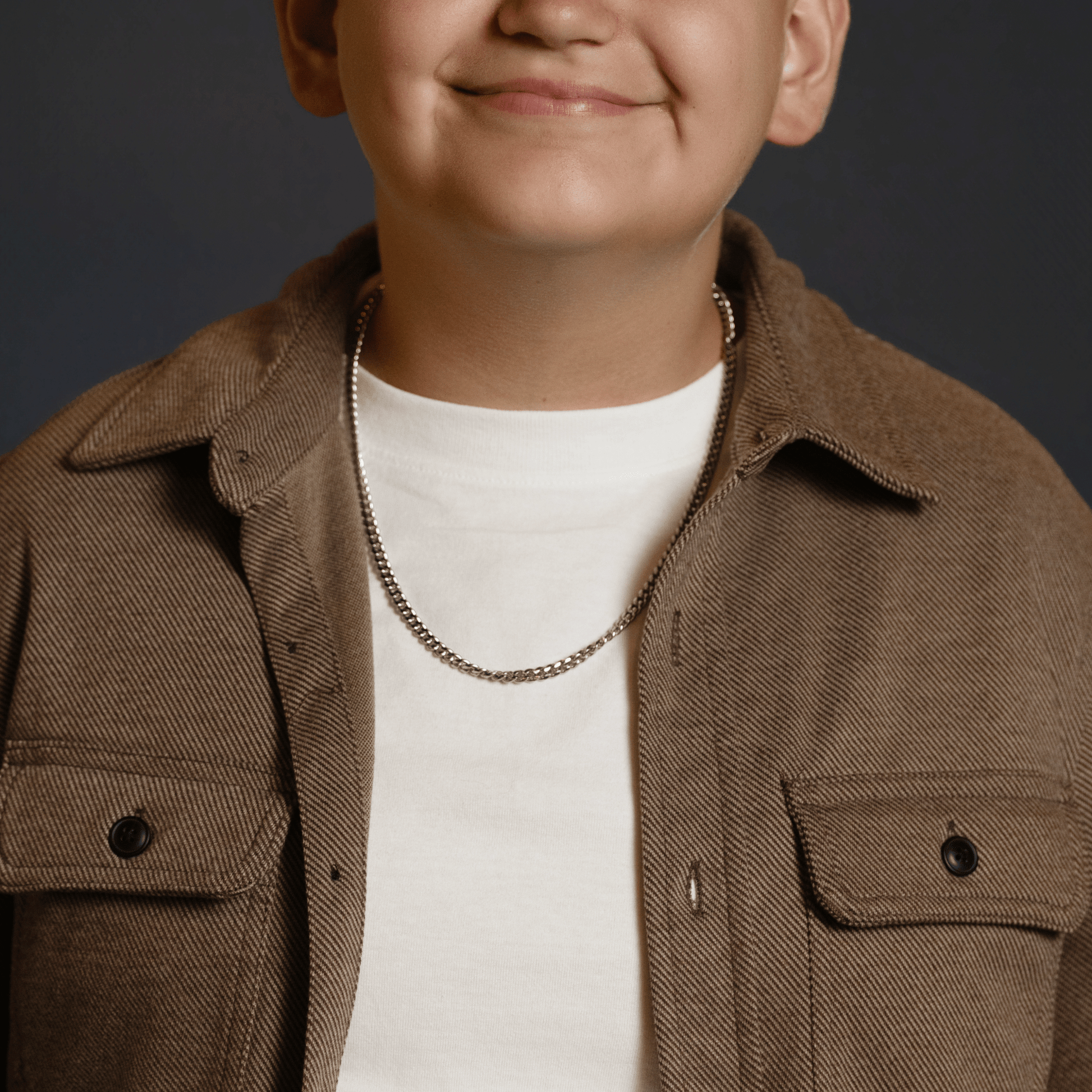 A close-up of a person wearing a brown corduroy overshirt over a white t-shirt, accessorized with a silver chain necklace, against a dark gray background.