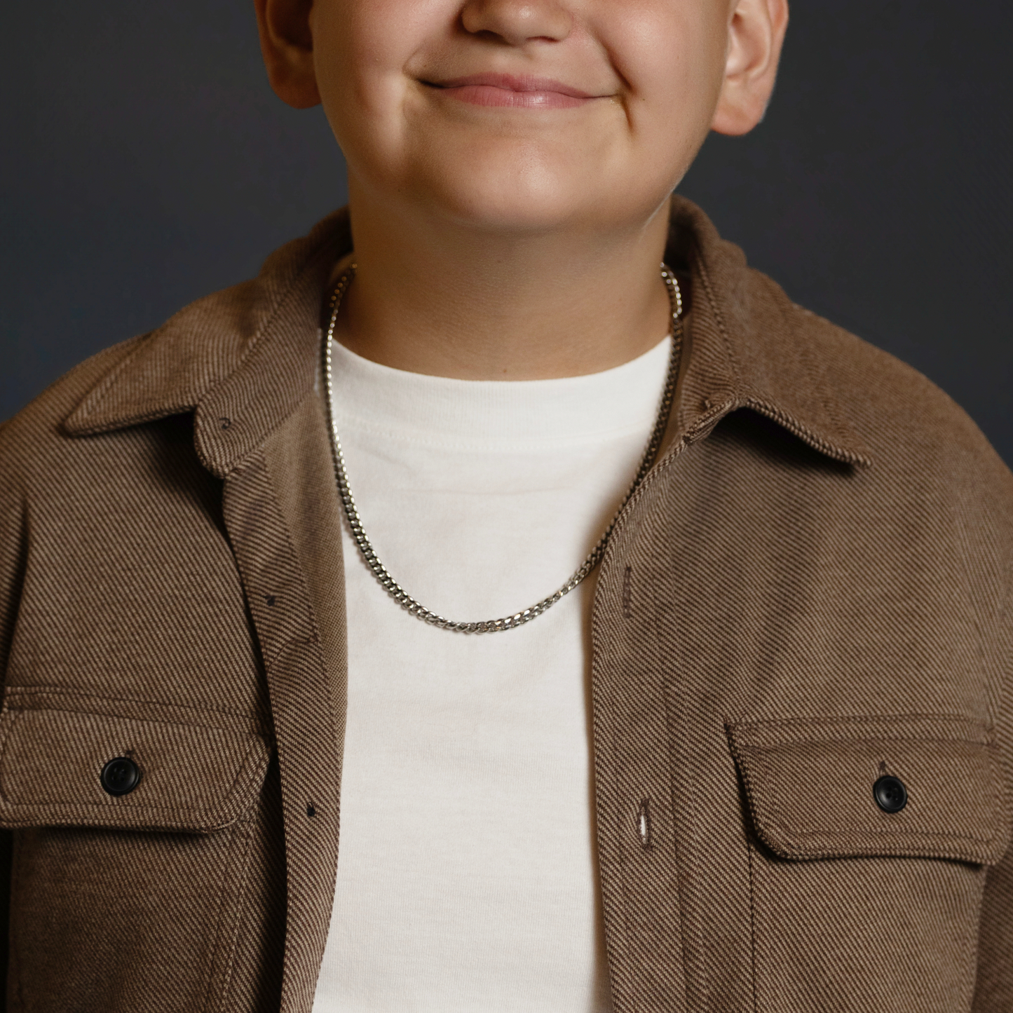 A close-up of a person wearing a brown corduroy overshirt over a white t-shirt, accessorized with a silver chain necklace, against a dark gray background.