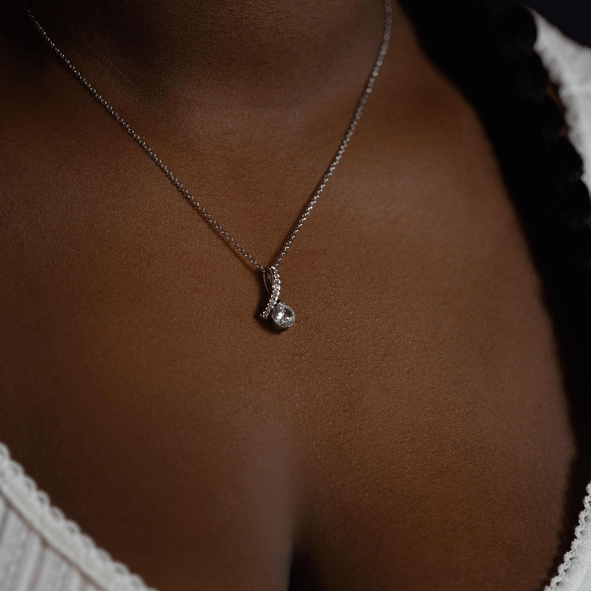 person wearing a silver chain necklace with a small silver pendant featuring a clear gemstone, shown against dark brown skin