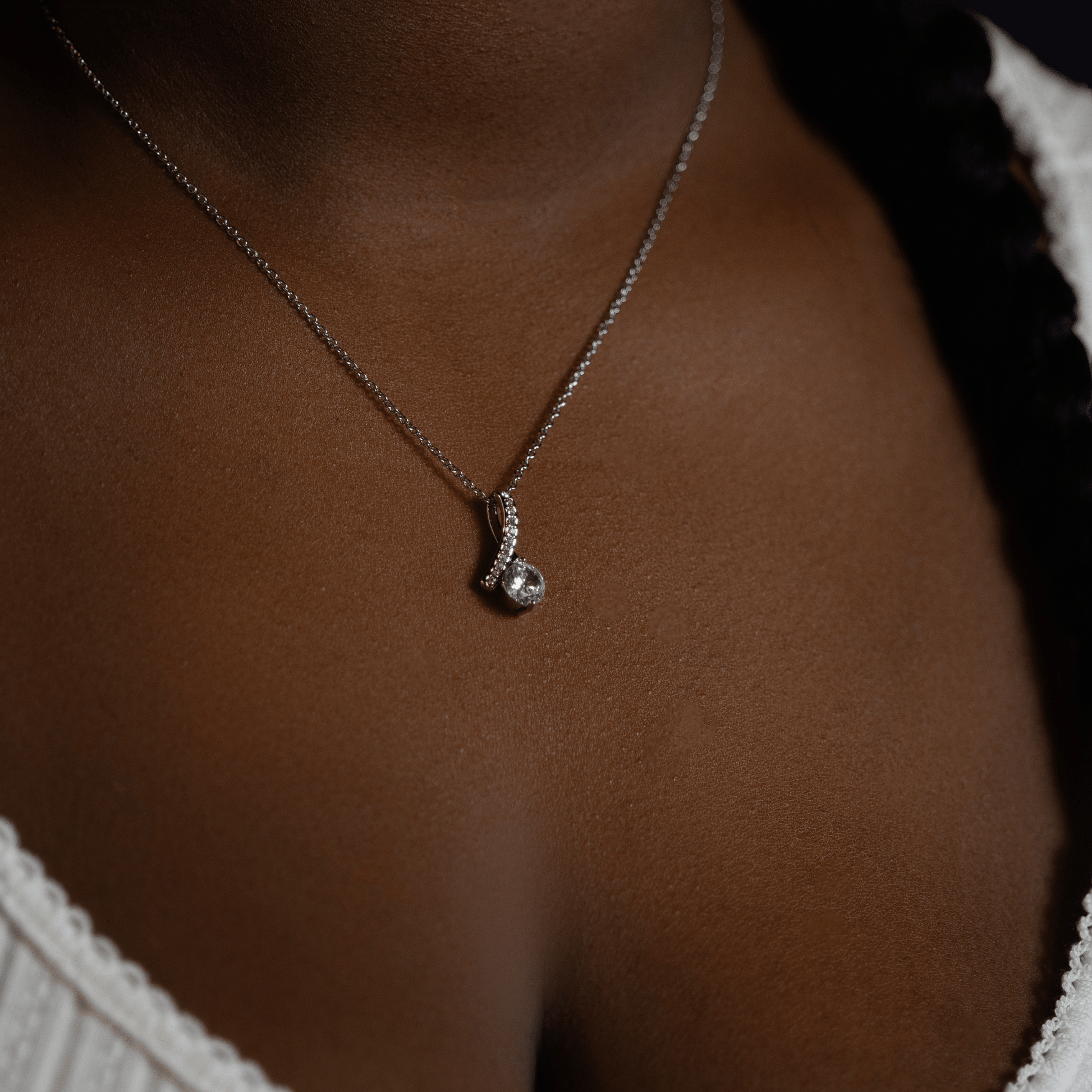 person wearing a silver chain necklace with a small silver pendant featuring a clear gemstone, shown against dark brown skin