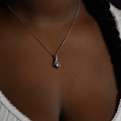 person wearing a silver chain necklace with a small silver pendant featuring a clear gemstone, shown against dark brown skin
