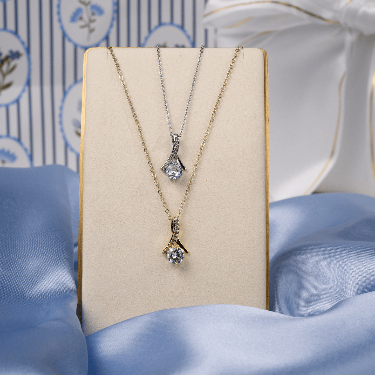 Two pendant necklaces displayed on a cream jewelry stand: one gold-tone and one silver-tone, each with a clear gemstone