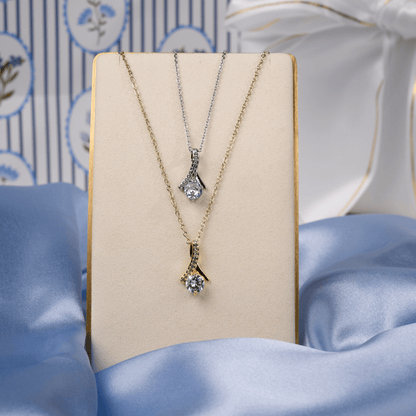 Two pendant necklaces displayed on a cream jewelry stand: one gold-tone and one silver-tone, each with a clear gemstone